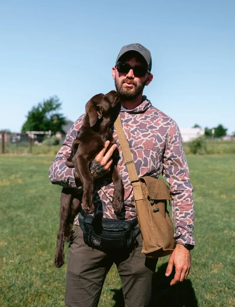 Justin Rie, Master Trainer for Off Leash K9 Training Asheville, holding a chocolate Labrador puppy in a training field